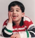 School photo of a boy with dark hair and pale skin, wearing a red, green, white, and black-striped shirt. He is missing his upper front teeth. He has his head propped on one hand. His eyes are half-closed.