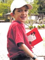 Photo of a boy in a red shirt and white baseball cap; he has light skin and dark hair, and is holding a book.