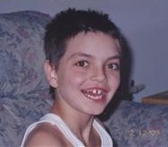 Photo of Kyle Potts, a young boy with short dark hair, brown eyes, and tanned skin, wearing a white tank top. His wide smile reveals a gap between his front teeth.