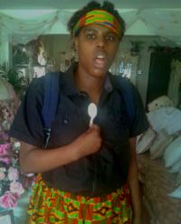 Photo of an African-American young woman wearing a black shirt, colorful skirt and matching headband. She is wearing a backpack and holding a spoon. Her mouth is open.