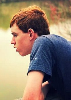 Photo of a teenage boy with auburn hair and fair skin, wearing a gray T-shirt. He is in profile, gazing across the water of a pond. His expression is neutral, with his mouth slightly open.