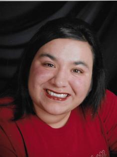 Photo: A heavyset woman wearing a red sweater and red lipstick. Her chin-length straight black hair contrasts with her fair skin.