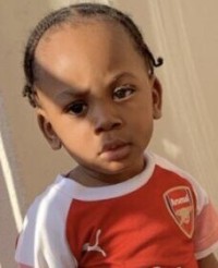 Photo of a toddler with his hair in small braids; he has light-brown skin and he is wearing a red and white sports jersey. His expression is thoughtful and somewhat puzzled.