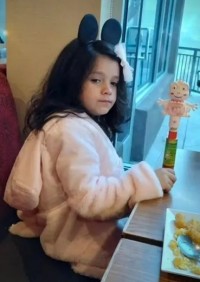 Photo of a young girl sitting at a table, wearing a fluffy pink robe. She has light skin and long black hair with a pink bow in it and a mouse-ears headband.